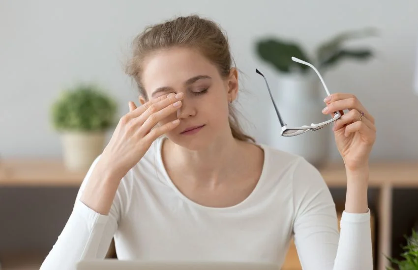 Mujer cansada frotándose los ojos al despertar, con signos de fatiga matutina y gafas en la mano, representando la falta de descanso reparador.